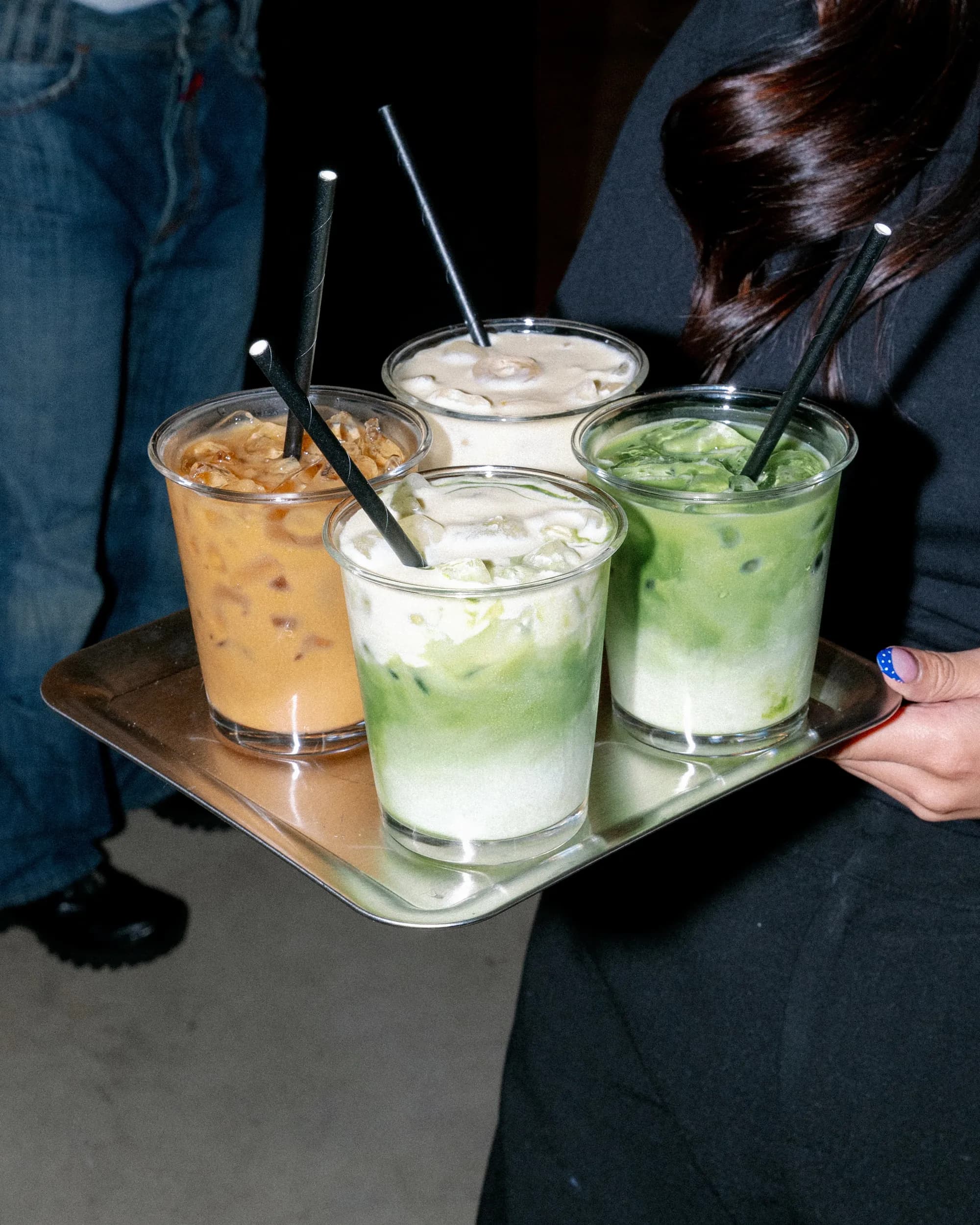 A steel tray carried by hand holding four iced drinks — an iced coffee, two matcha lattes and a cream-topped coffee — each with a black paper straw.