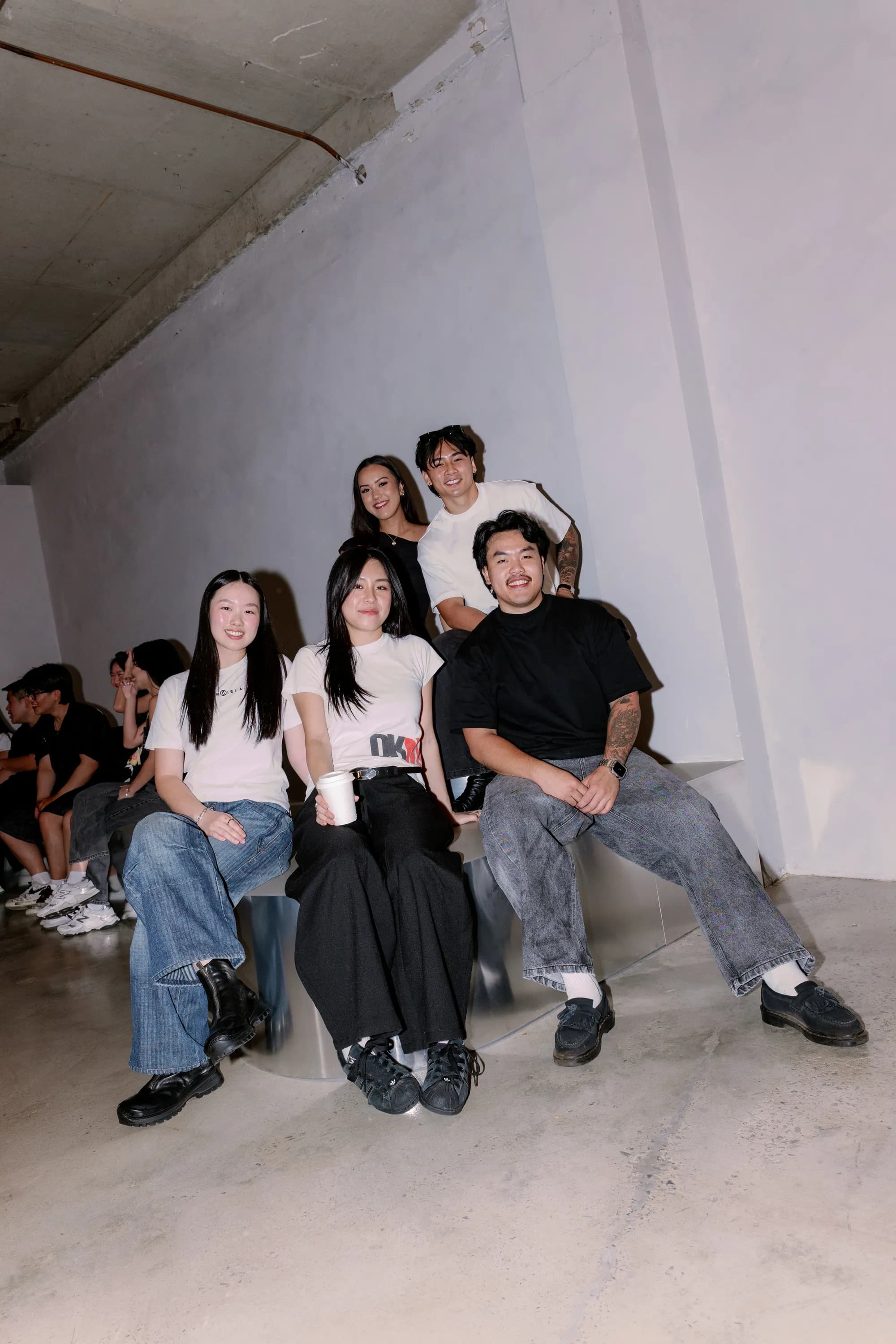 A group of five sitting together on the polished steel bench at a Revival Studios event, with more of the crowd along the wall behind them.